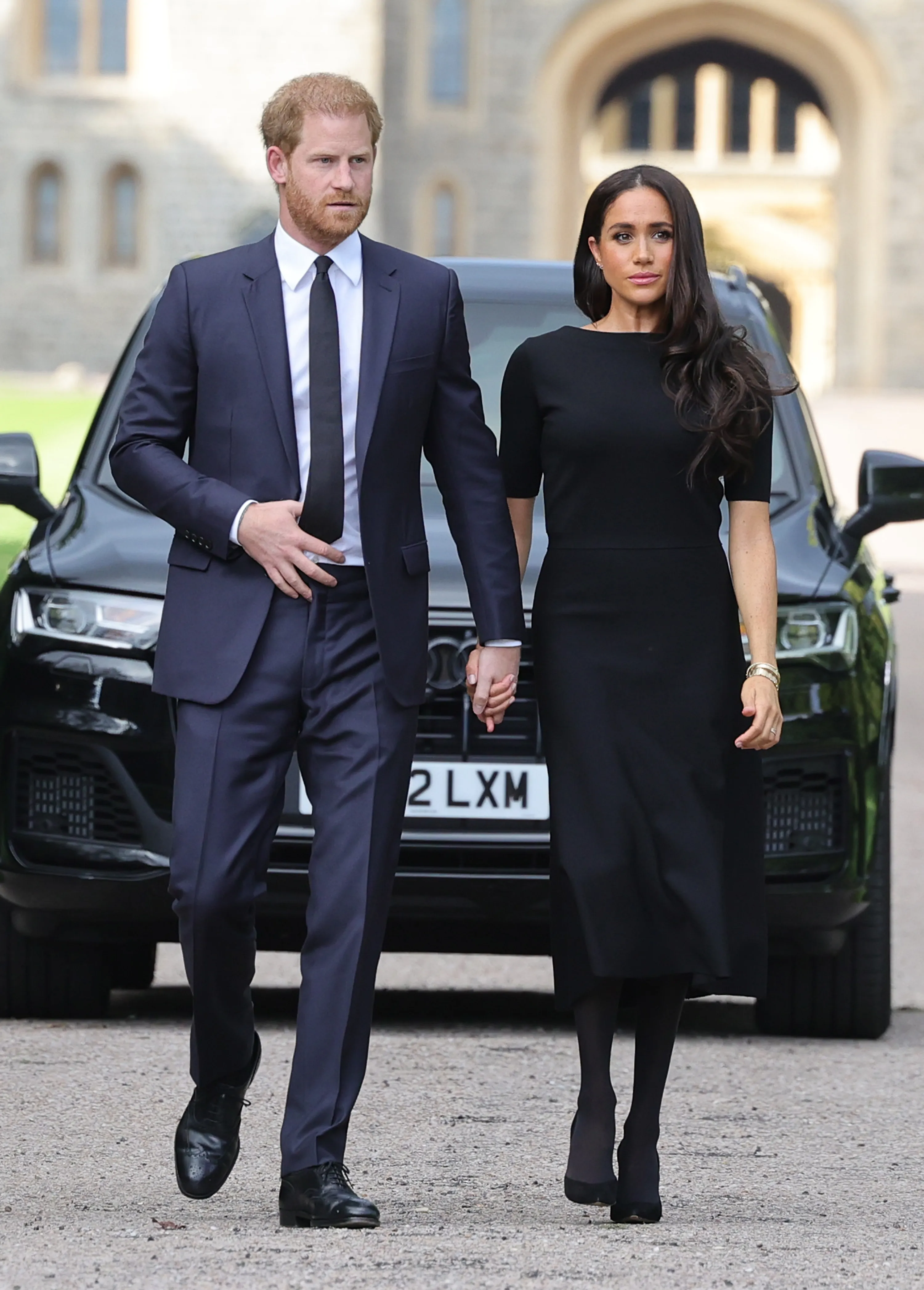 Prince Harry and Meghan Markle holding hands while walking, with a black car and Windsor Castle in the background.