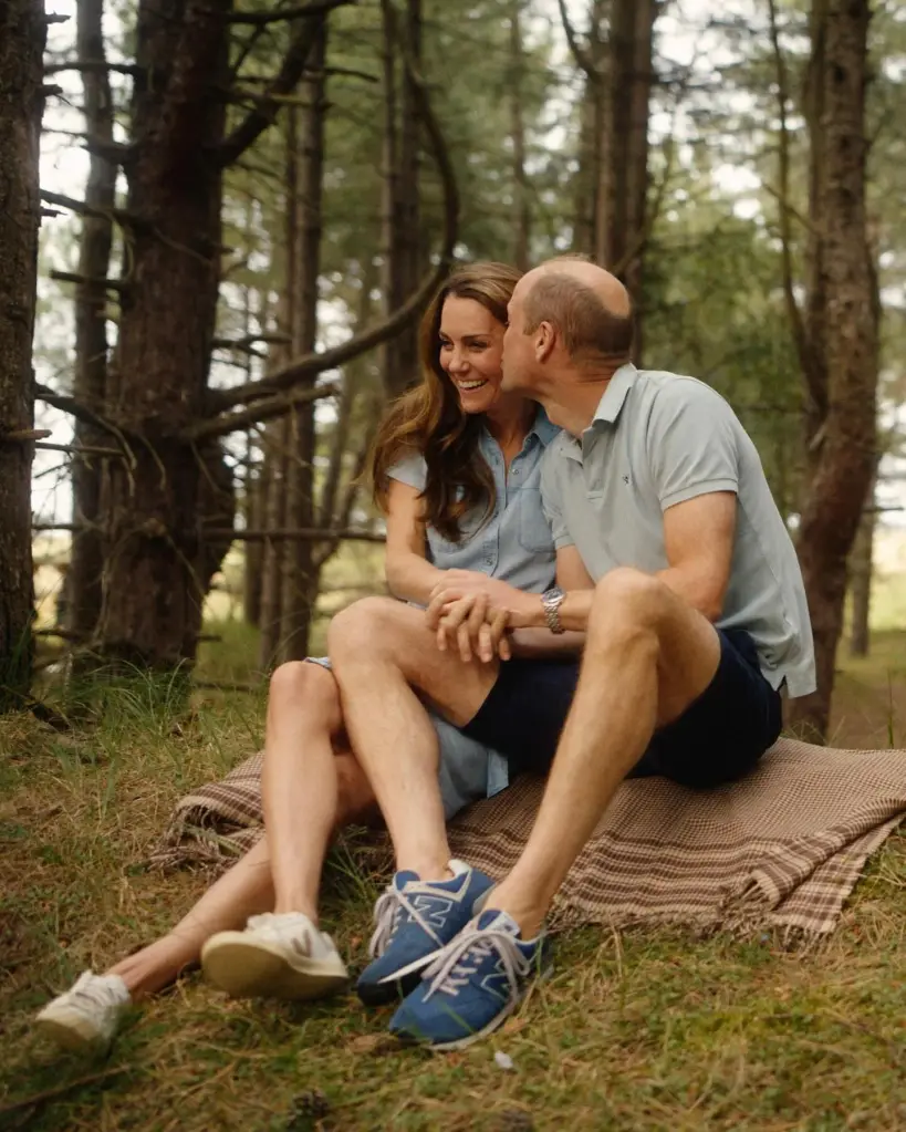 Prince William kissing Kate Middleton on the cheek while sitting on a blanket in a forest.