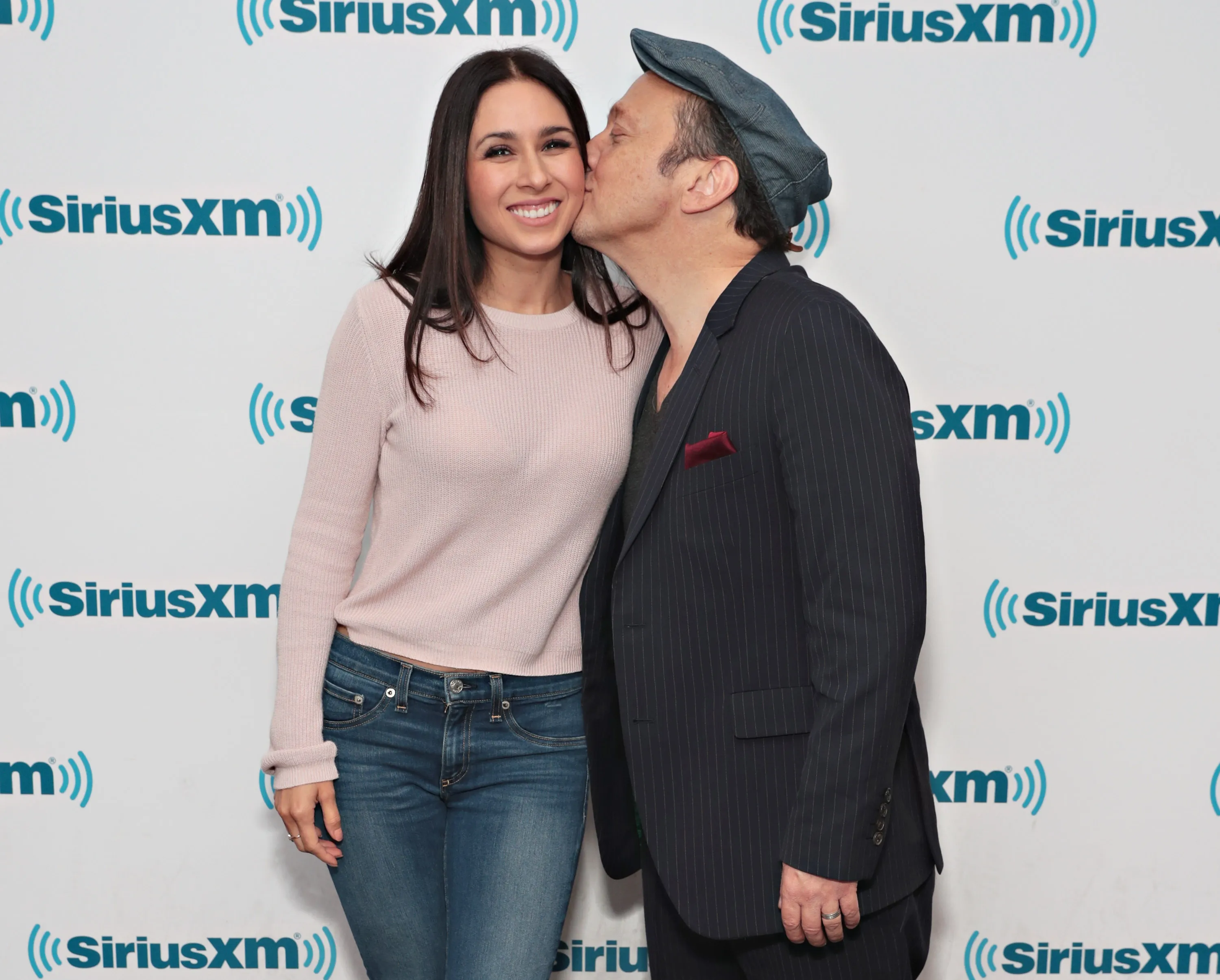 Producer Patricia Maya Schneider, smiling, is kissed on the cheek by actor Rob Schneider, in front of a white wall with the SiriusXM logo.