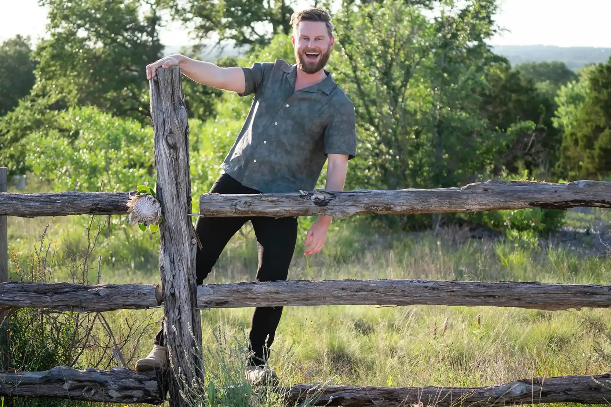 Bobby Berk behind a rustic wooden fence, with a protea flower attached to a fence post.