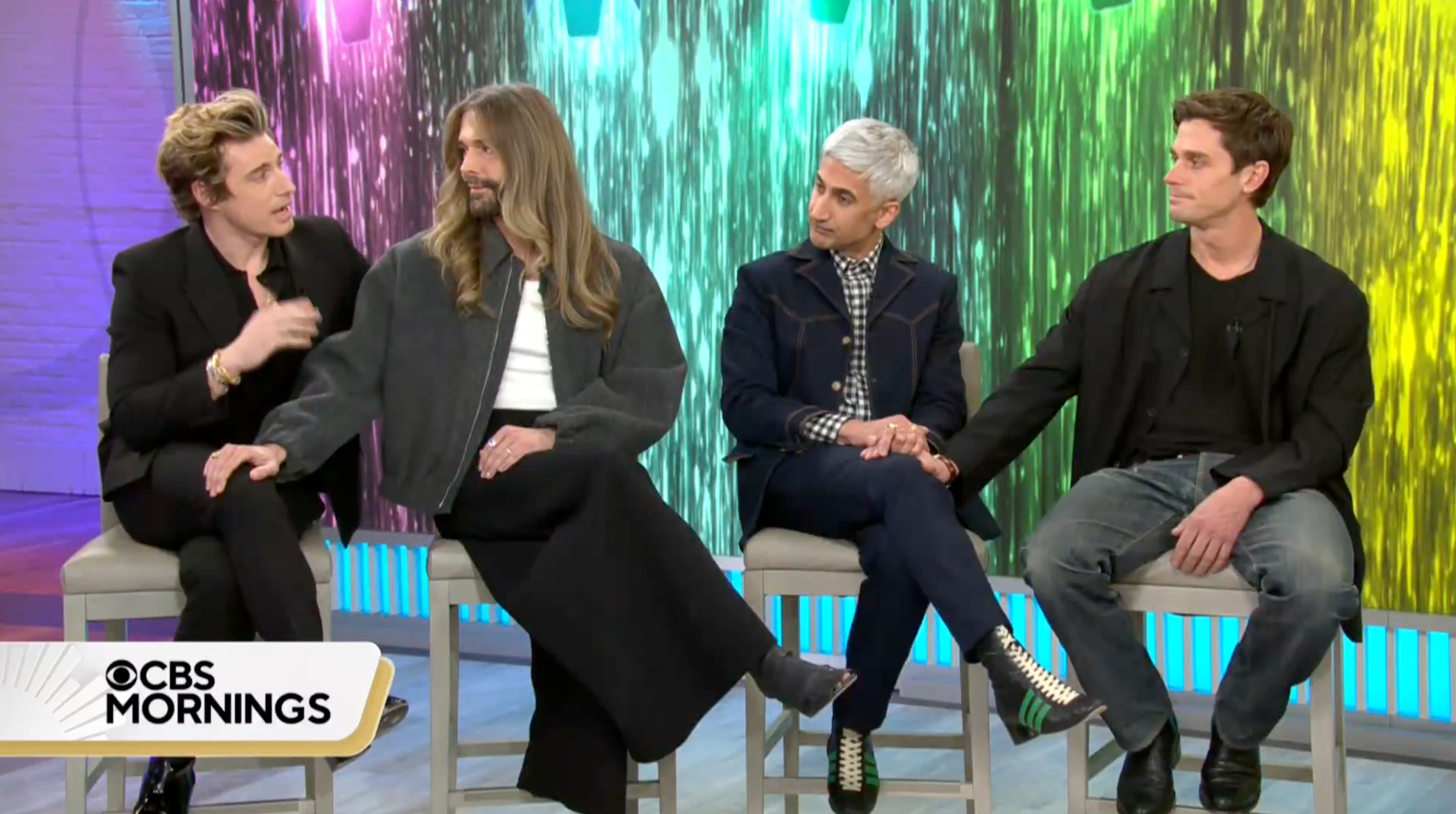 Four men, two of them members of the cast of Queer Eye, sit on stools in front of a colorful backdrop that shifts from pink to blue to green to yellow.