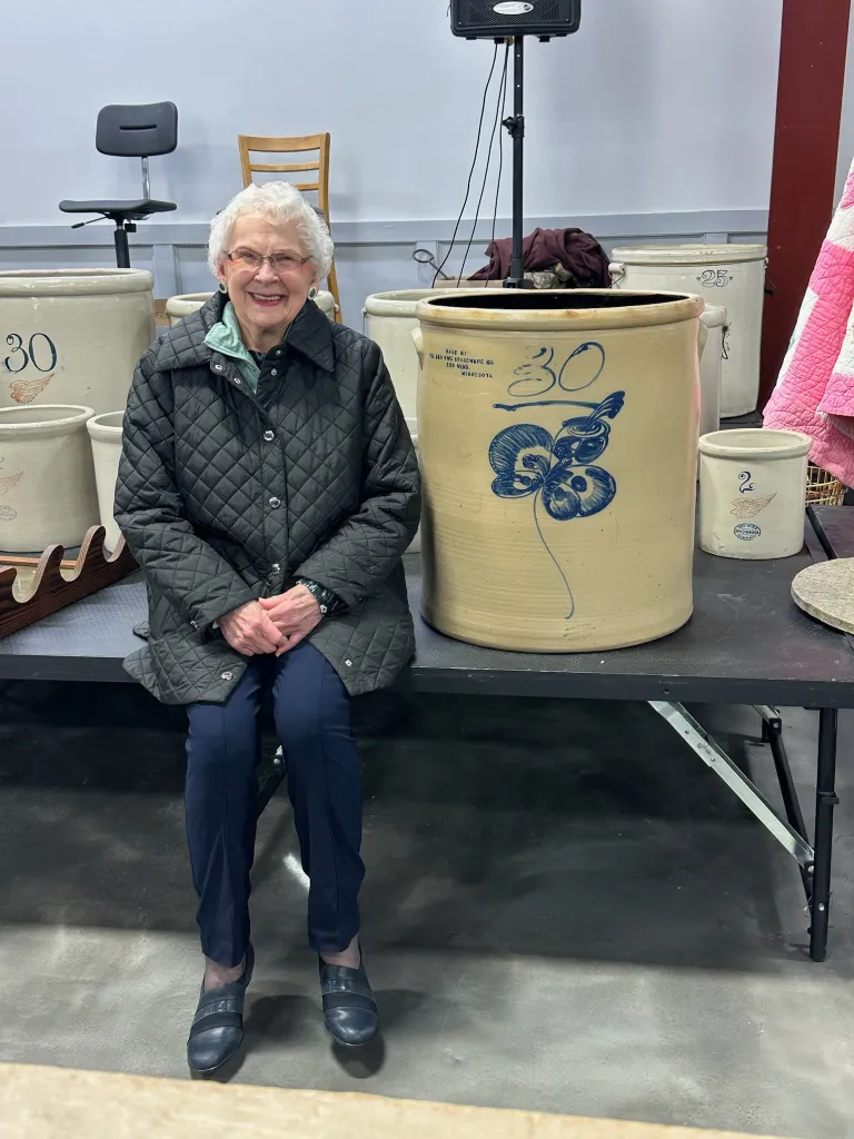 An elderly woman sits next to a large crock with 