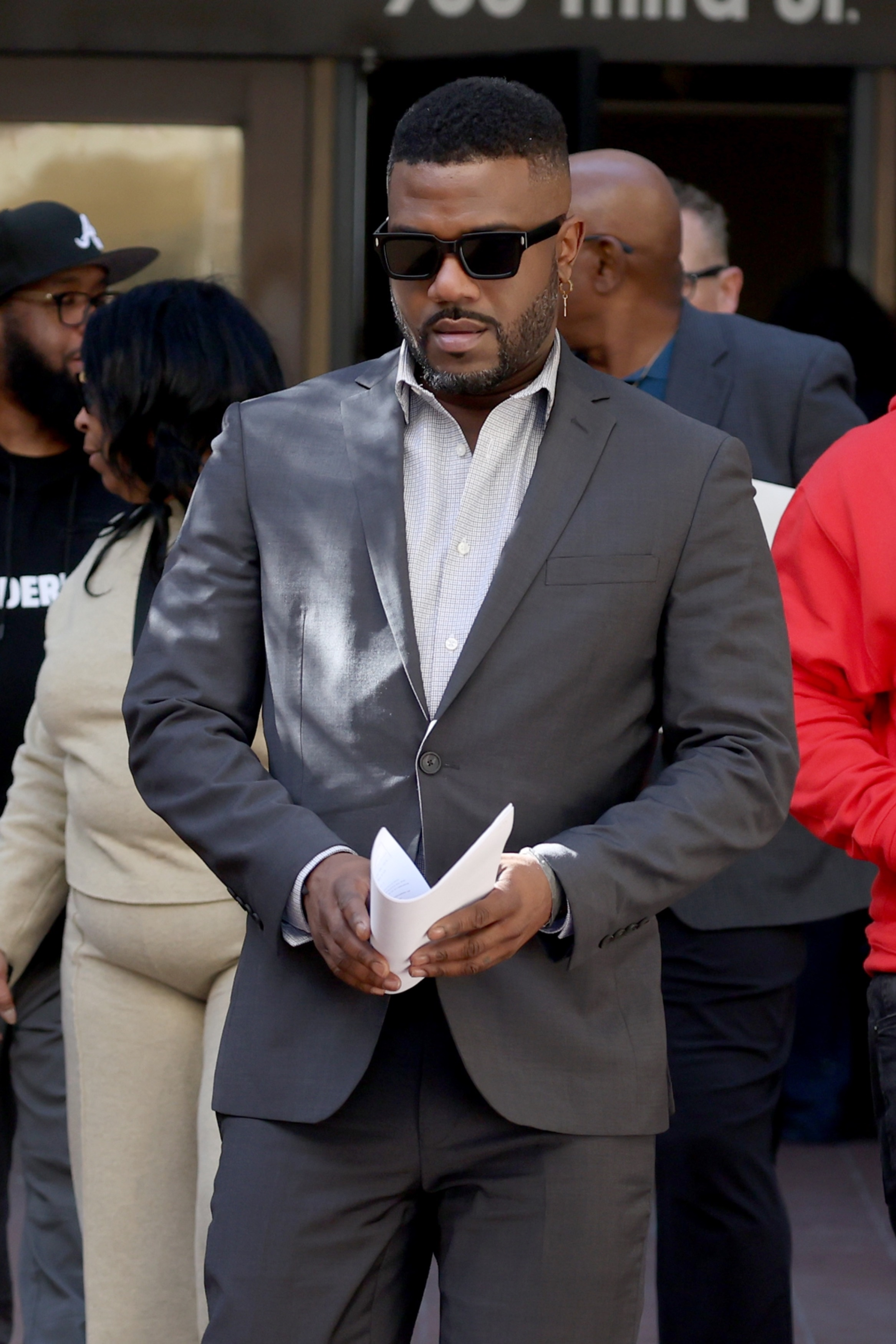 Ray J in a grey suit and sunglasses, holding papers, after a court hearing.