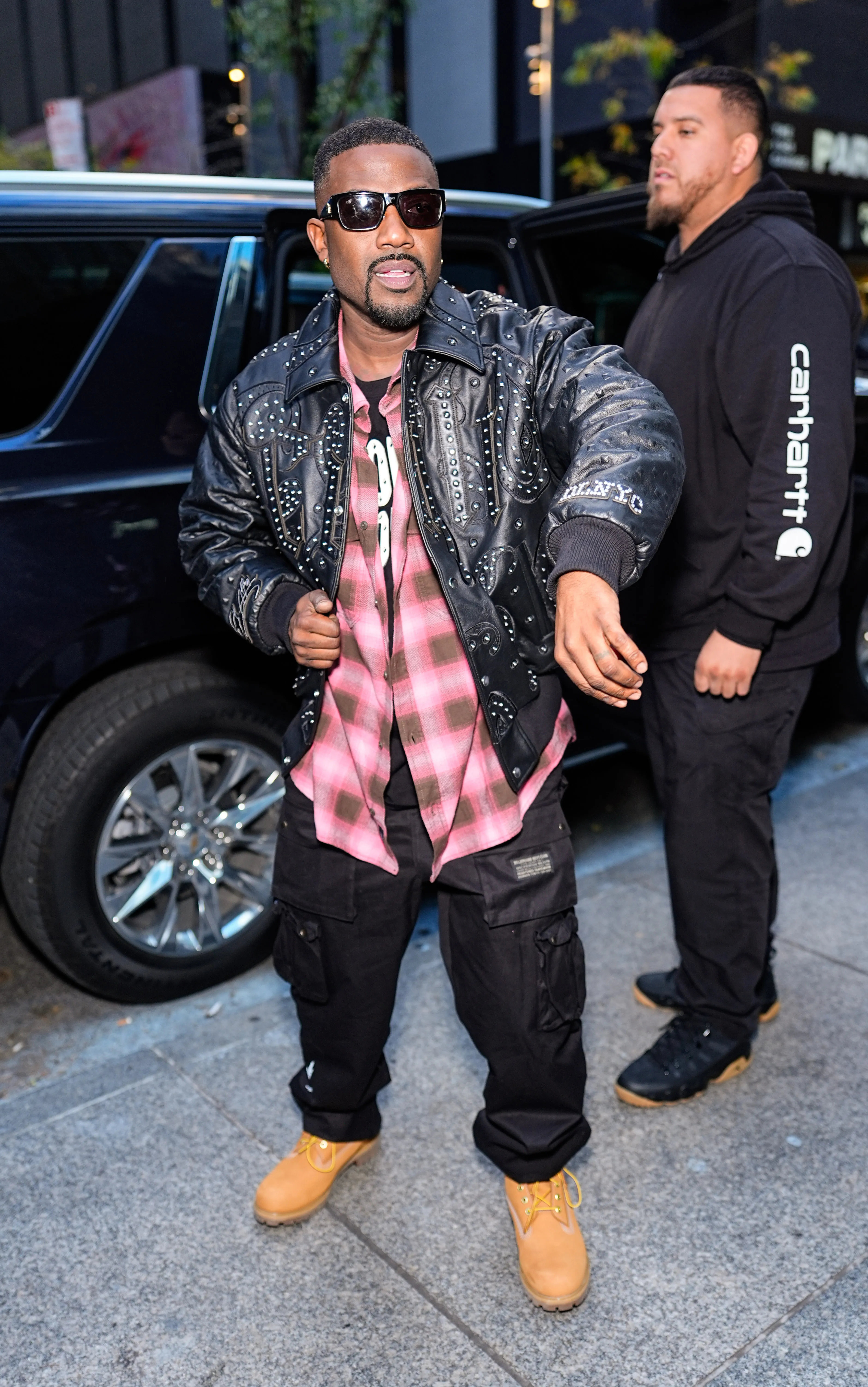 Ray J in a black studded leather jacket, pink plaid shirt, black cargo pants, and tan boots, standing next to a car.