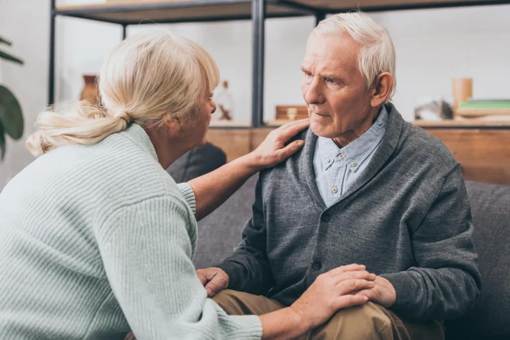 A retired woman touching her husband's shoulder as they both look at each other with sad expressions.