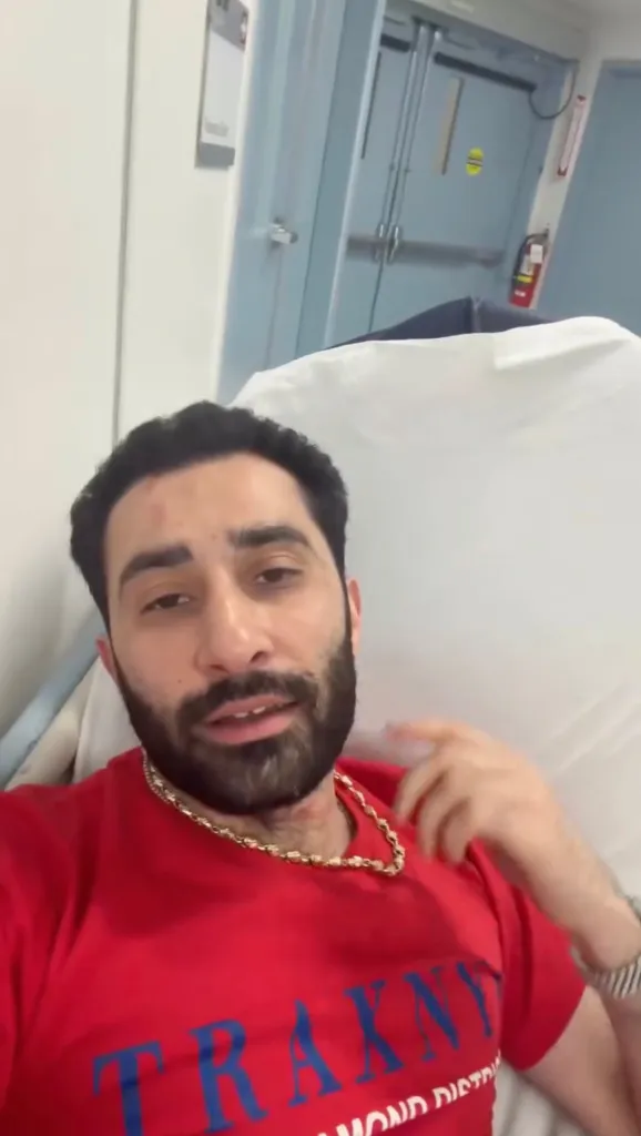 Man in red shirt and gold chain in a hospital bed with injuries to his forehead and neck.