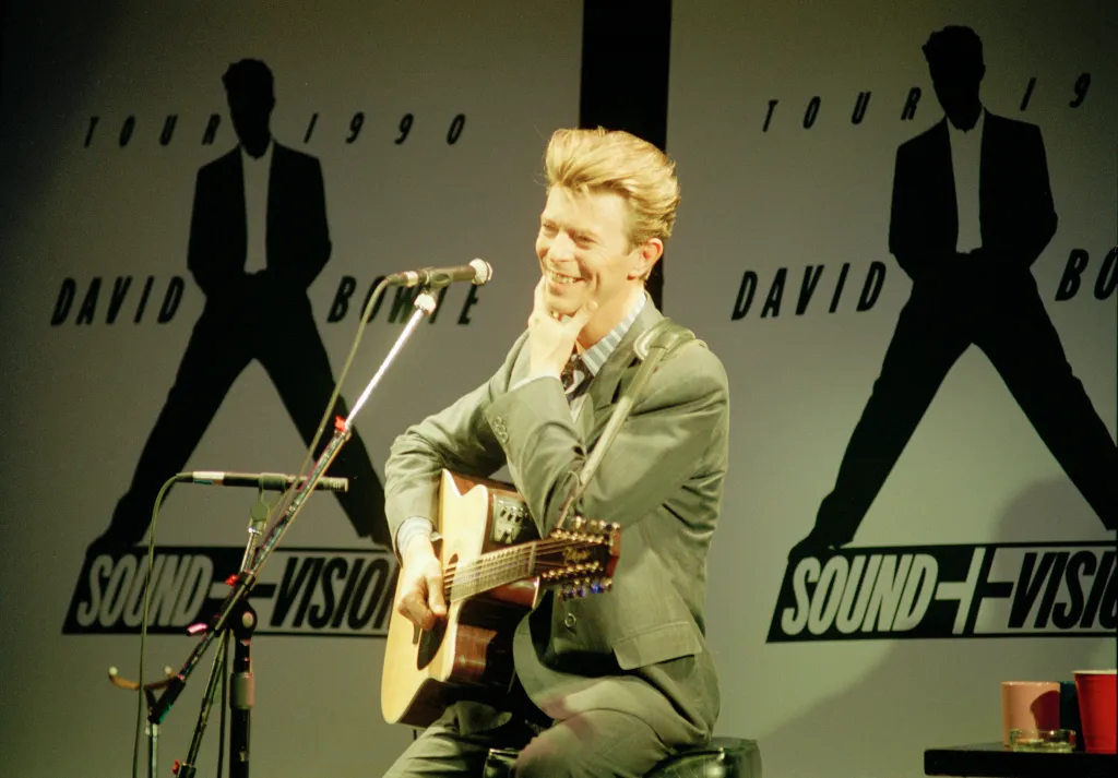 David Bowie playing an acoustic guitar and smiling at a press conference for his 1990 