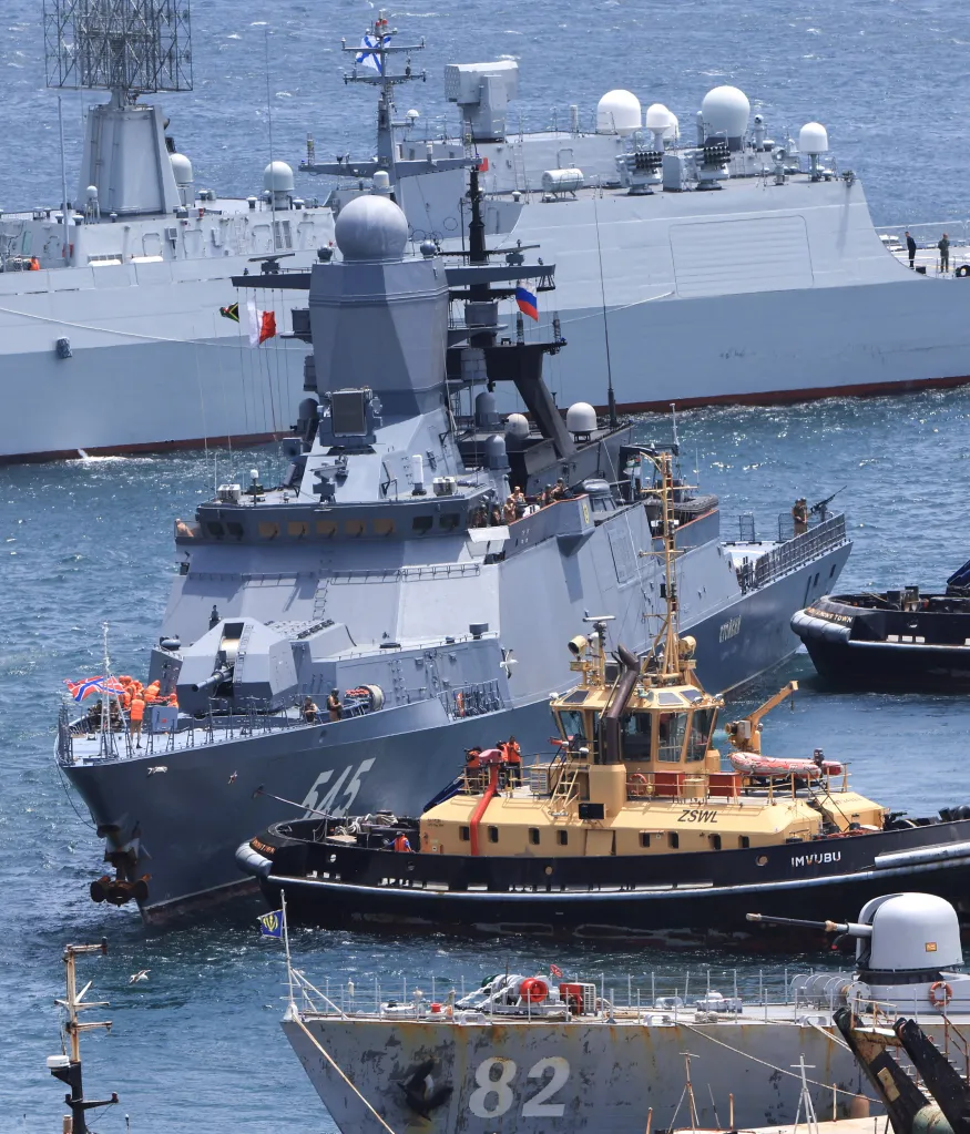 A Russian warship, with several people on deck, being guided by a tugboat in Cape Town, South Africa.