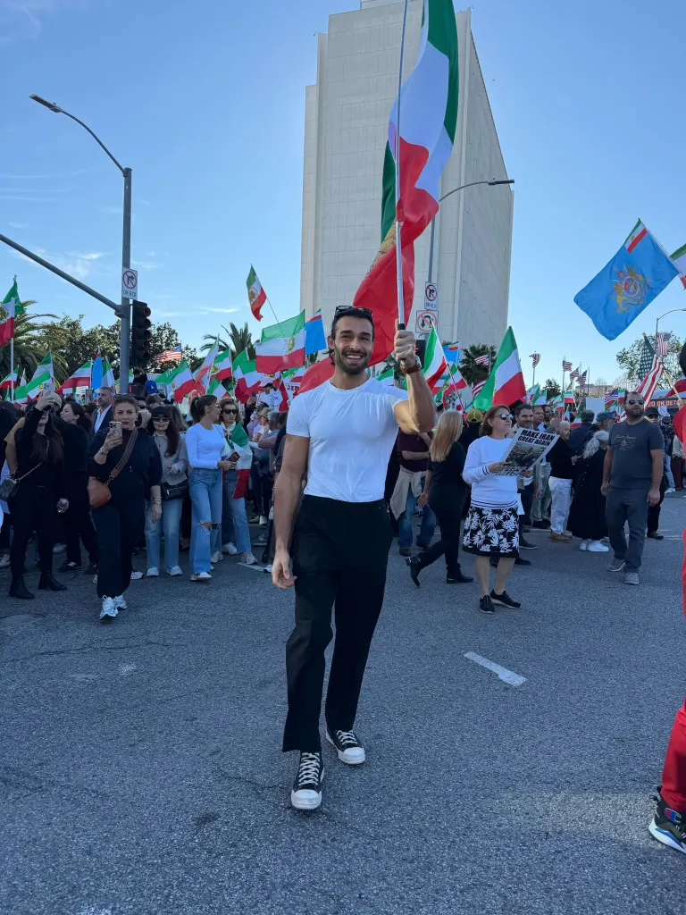 Sam Asghari protests in Los Angeles, holding an Iranian flag and smiling, surrounded by other protestors also holding Iranian flags.