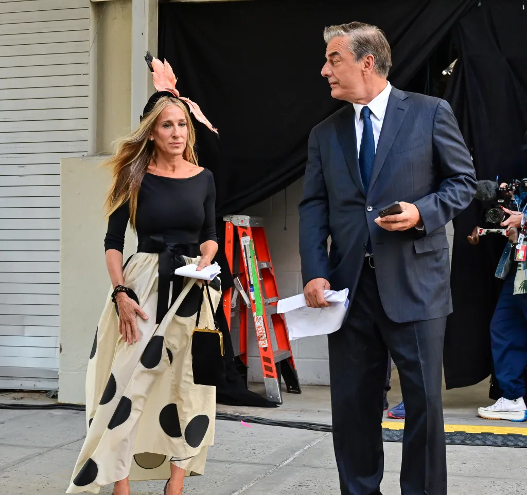 Sarah Jessica Parker in a polka dot dress and a feathery hat talks to Chris Noth in a blue suit.