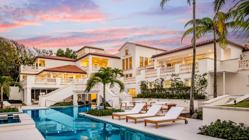 Luxury white mansion with red tile roof, palm trees, outdoor swimming pool, and lounge chairs at sunset.
