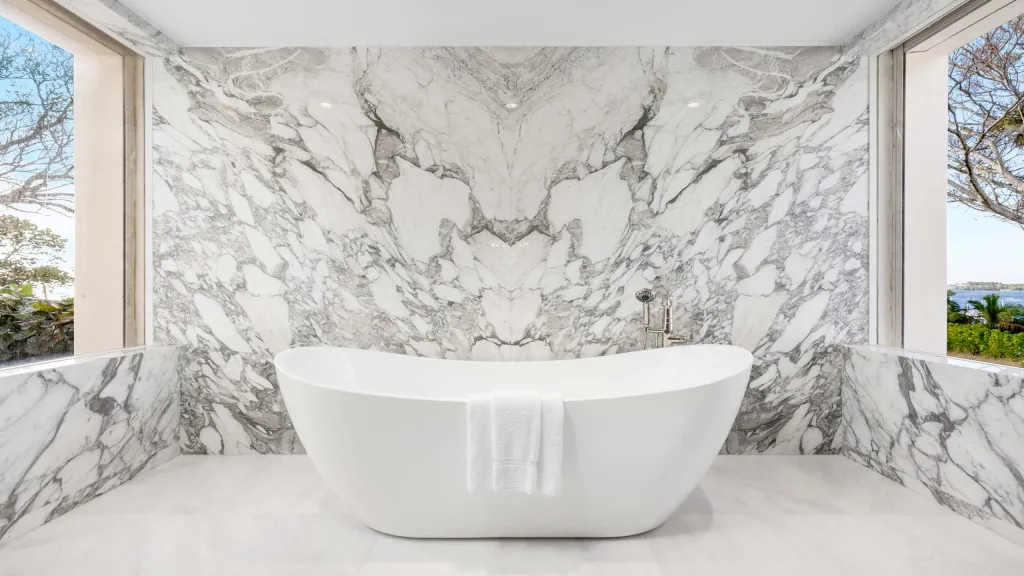 Bathroom in a Manalapan, Florida estate, with a white freestanding tub against a marble wall and large windows overlooking the water.