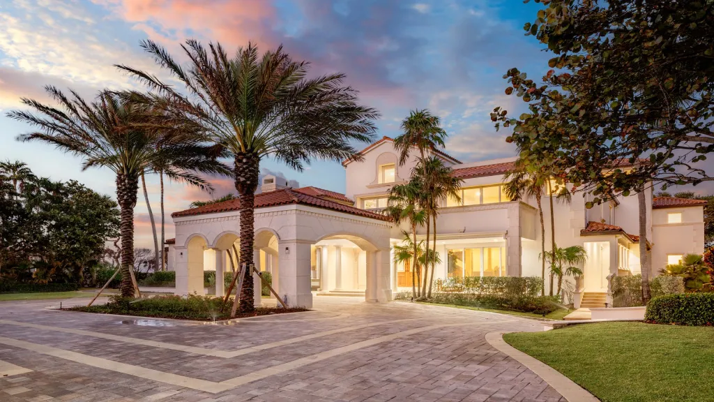 The Sean Hannity estate in Manalapan, Florida, with a curved driveway, arched portico, palm trees, and a sunset sky.