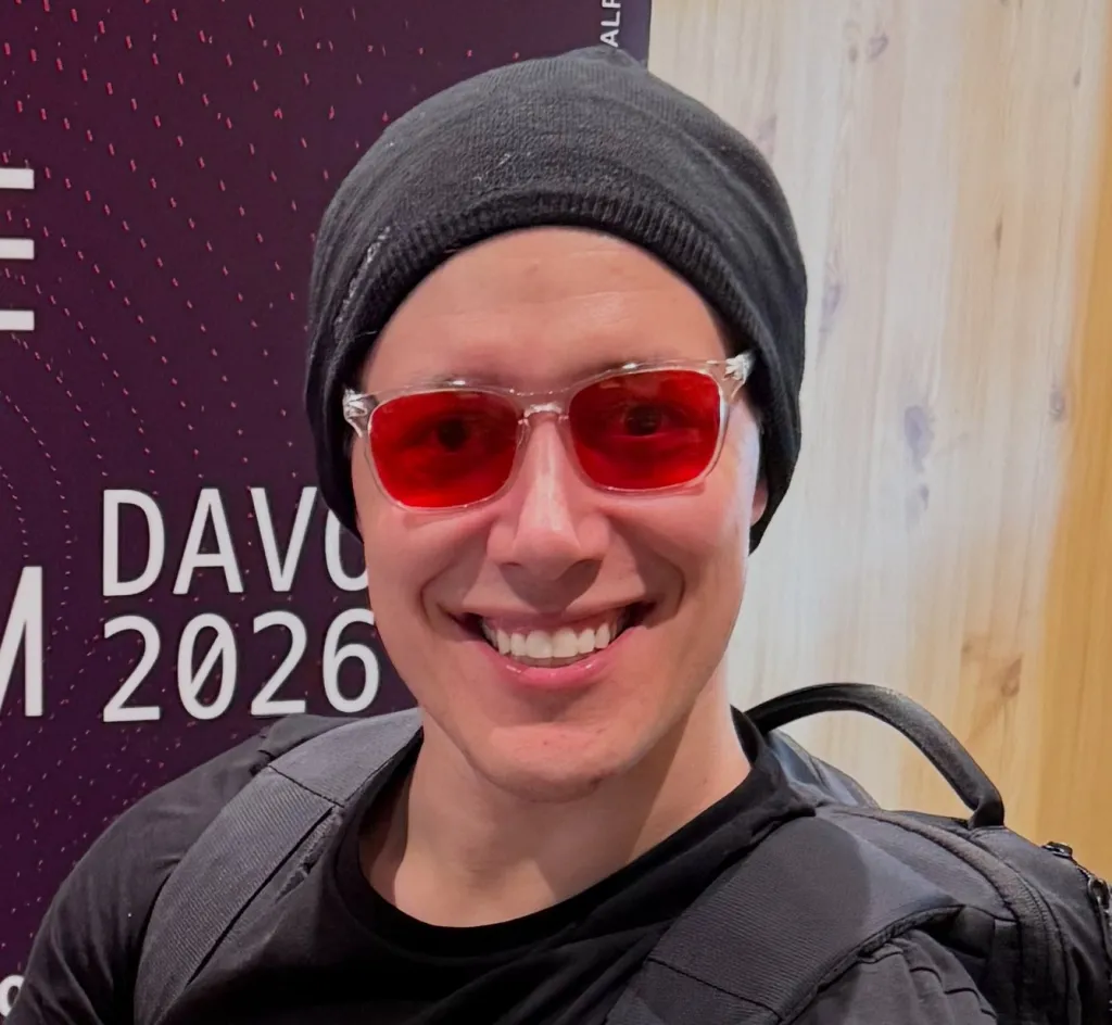 Sebastian Heyneman, smiling and wearing red sunglasses and a black beanie, against a purple backdrop with 
