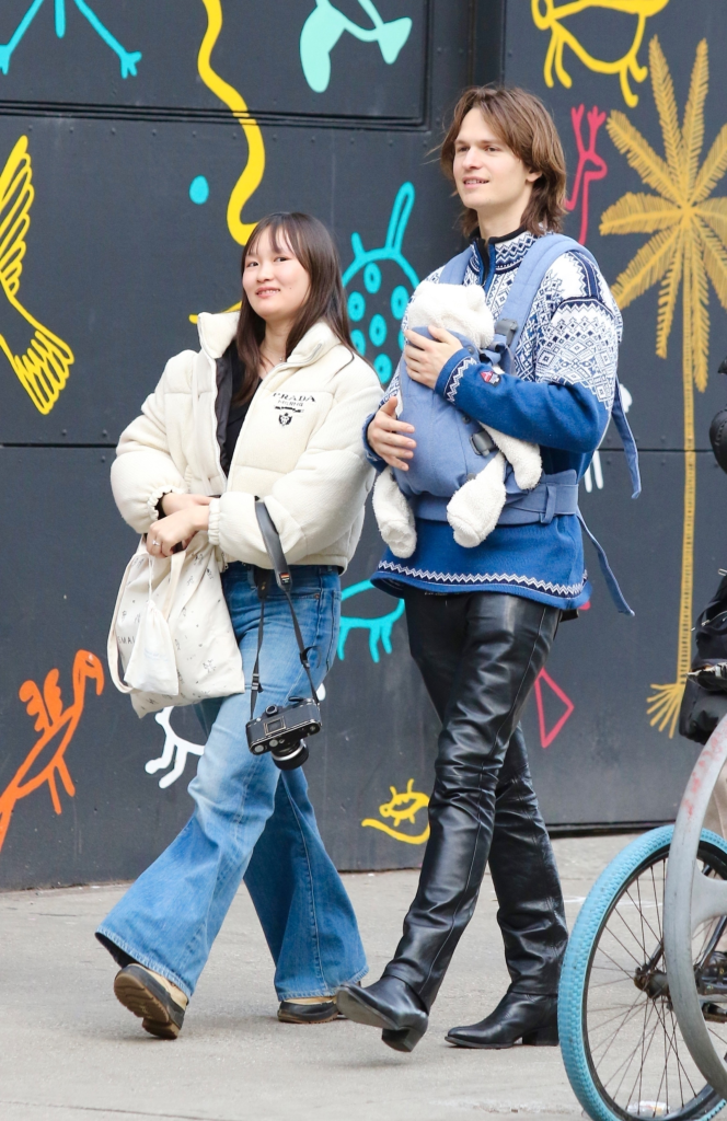 Ansel Elgort carries a baby in a papoose alongside his partner, outdoors in New York City.
