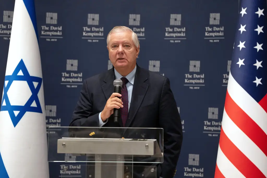 Senator Lindsey Graham speaks into a microphone at a podium, flanked by the Israeli and U.S. flags, with a backdrop displaying