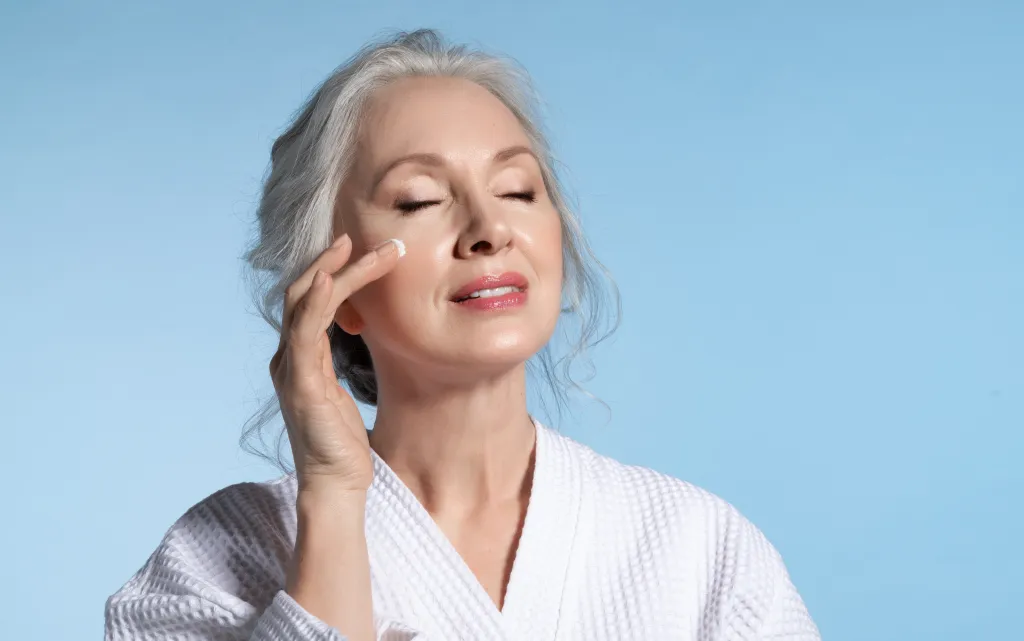 Senior woman with eyes closed applying moisturizing cream to her face.