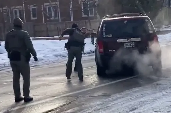 Two federal agents apprehending a person attempting to flee capture by ICE, with one agent holding the person near the back of a dark SUV emitting exhaust, on a snowy street.