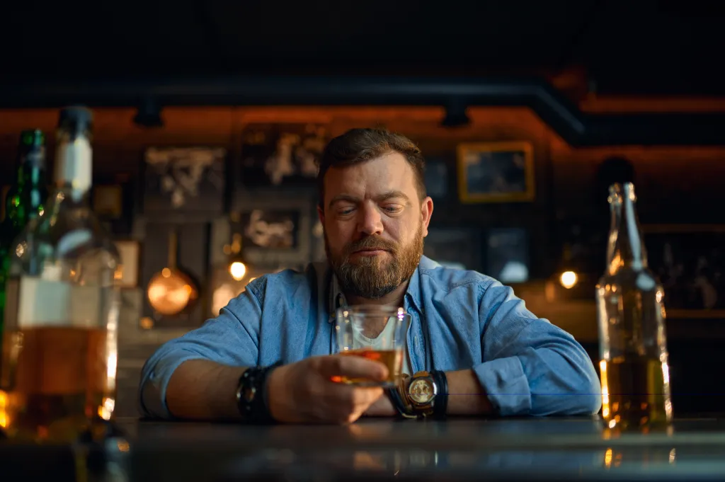 A serious, bearded man in a denim shirt sitting at a bar counter, holding a glass of alcohol and looking down.