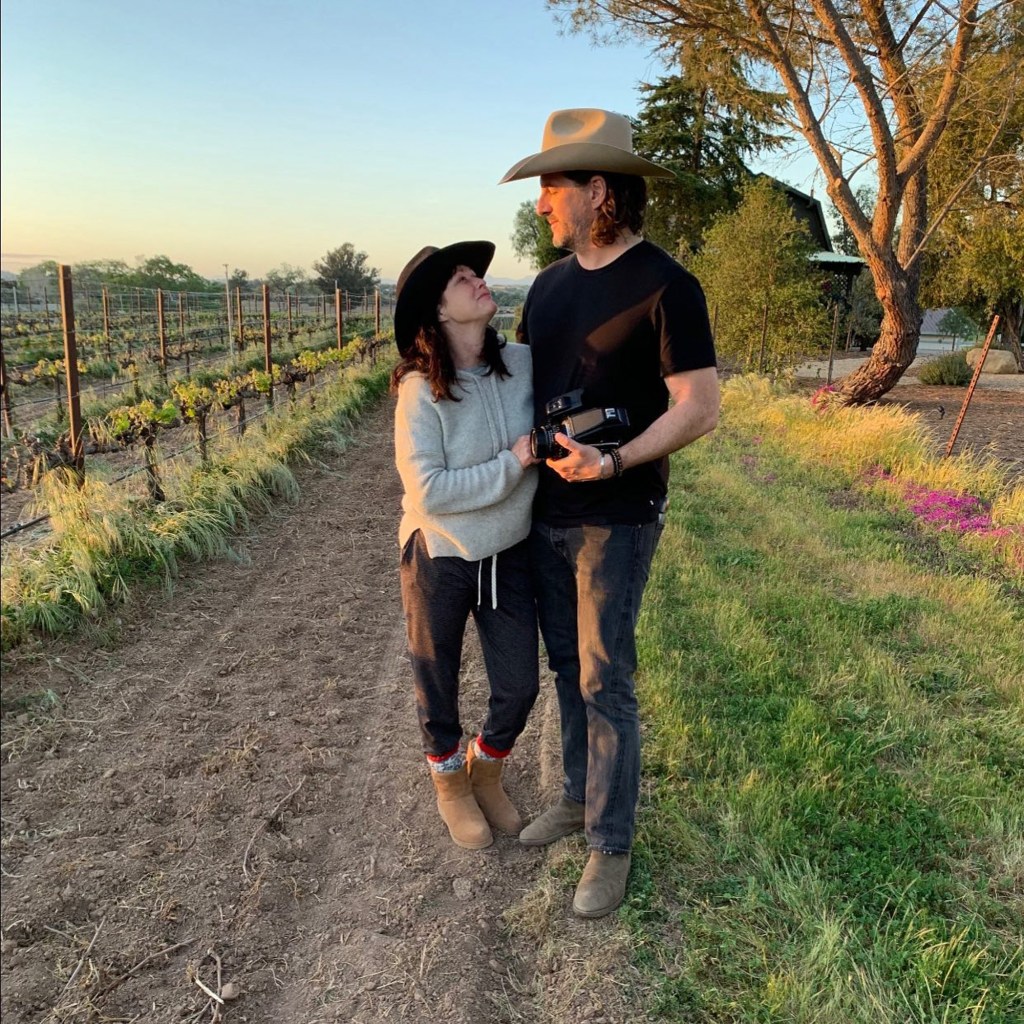 Shannen Doherty and Kurt Iswarienko in a vineyard, both wearing hats, looking at each other.
