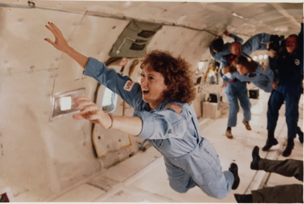 Christa McAuliffe, wearing a light blue flight suit, floats with arms outstretched in a zero-gravity training aircraft.