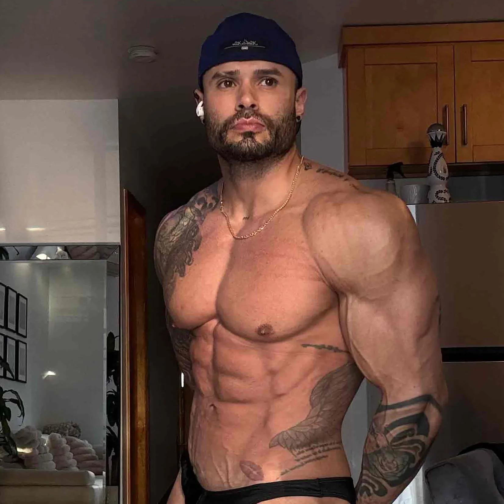 Christian Bueno, a bodybuilder, stands in a blue cap, black brief, with visible tattoos, showing his physique with an expression of concern.