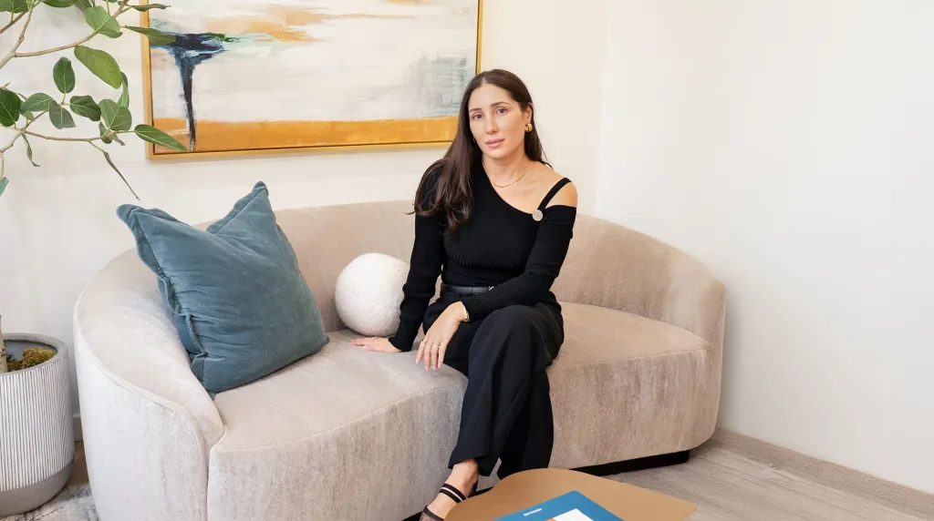 A woman in a black top and pants sits on a light brown couch