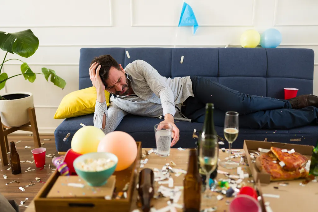 A man lies on a couch with a headache, surrounded by party debris like confetti, balloons, an empty pizza box, and bottles.