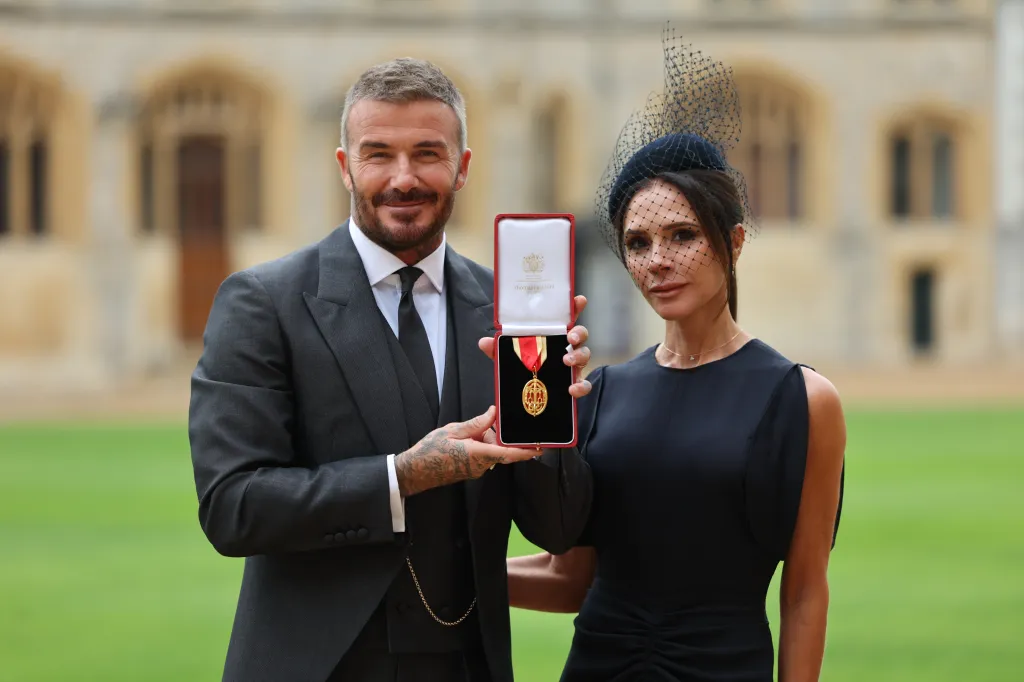 David Beckham, in a grey suit, white shirt and tie, and Victoria Beckham, in a navy blue dress and hat, holding up David's knighthood medal in a red box.