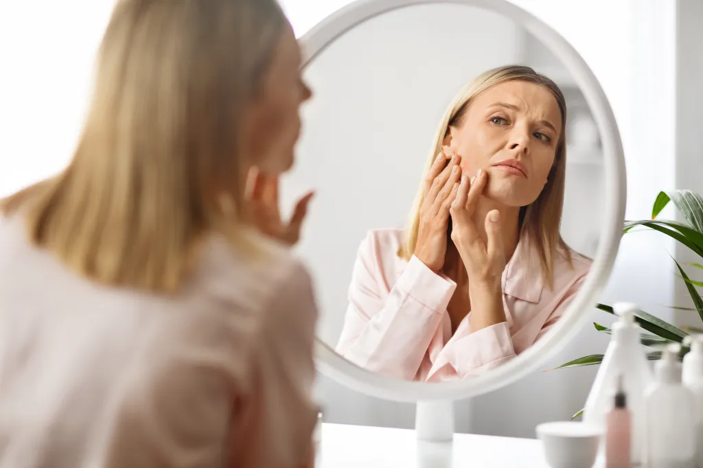 Upset middle-aged woman looking at pimples on her cheek in a mirror.