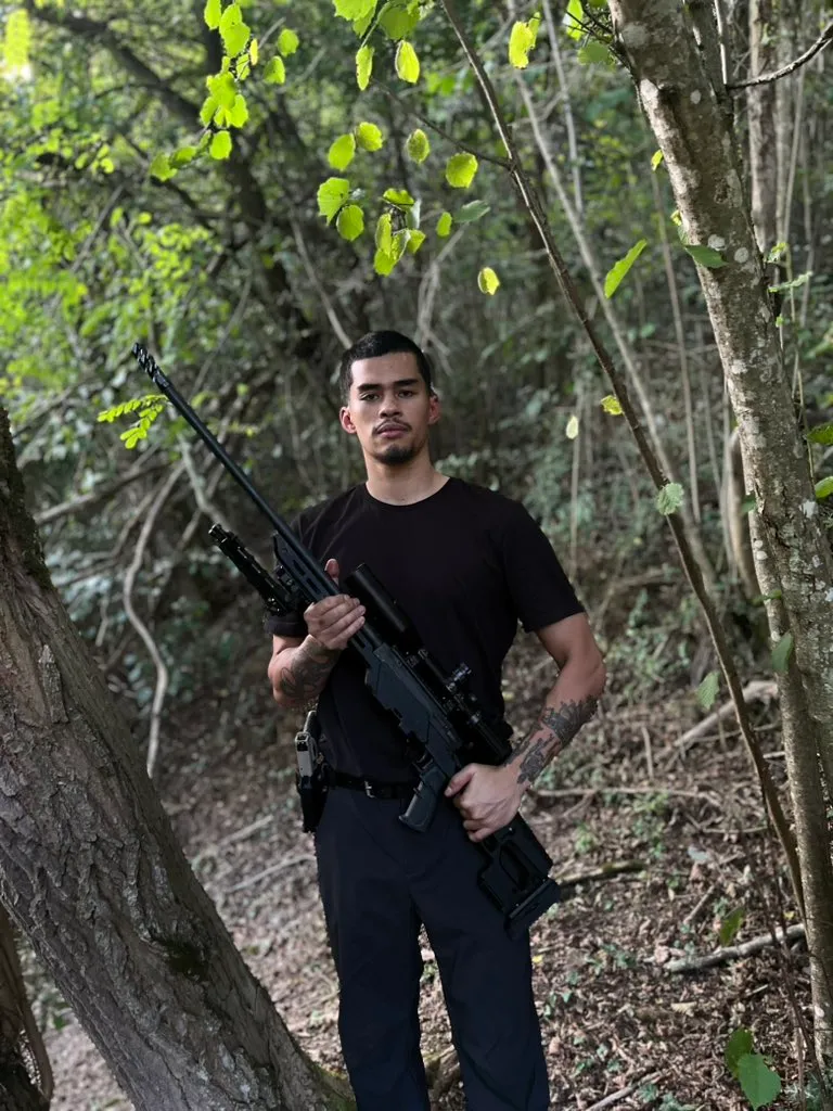 Sneako, also known as Nico Kenn De Balinthazy, holds a rifle in a wooded area.