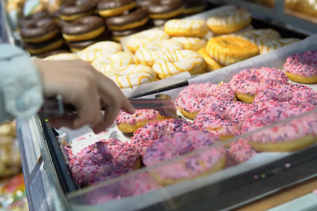 Hand using tongs to pick up a pink-frosted donut with sprinkles from a display case.
