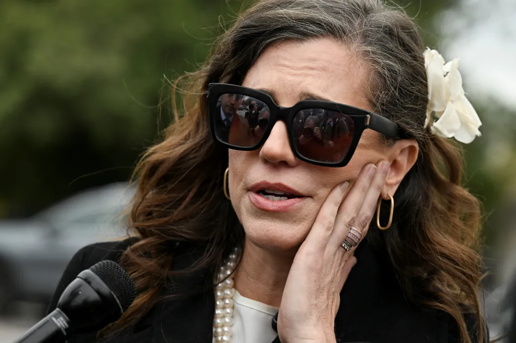 U.S. Representative Nancy Mace speaks to the media with a microphone, wearing sunglasses and a flower in her hair.