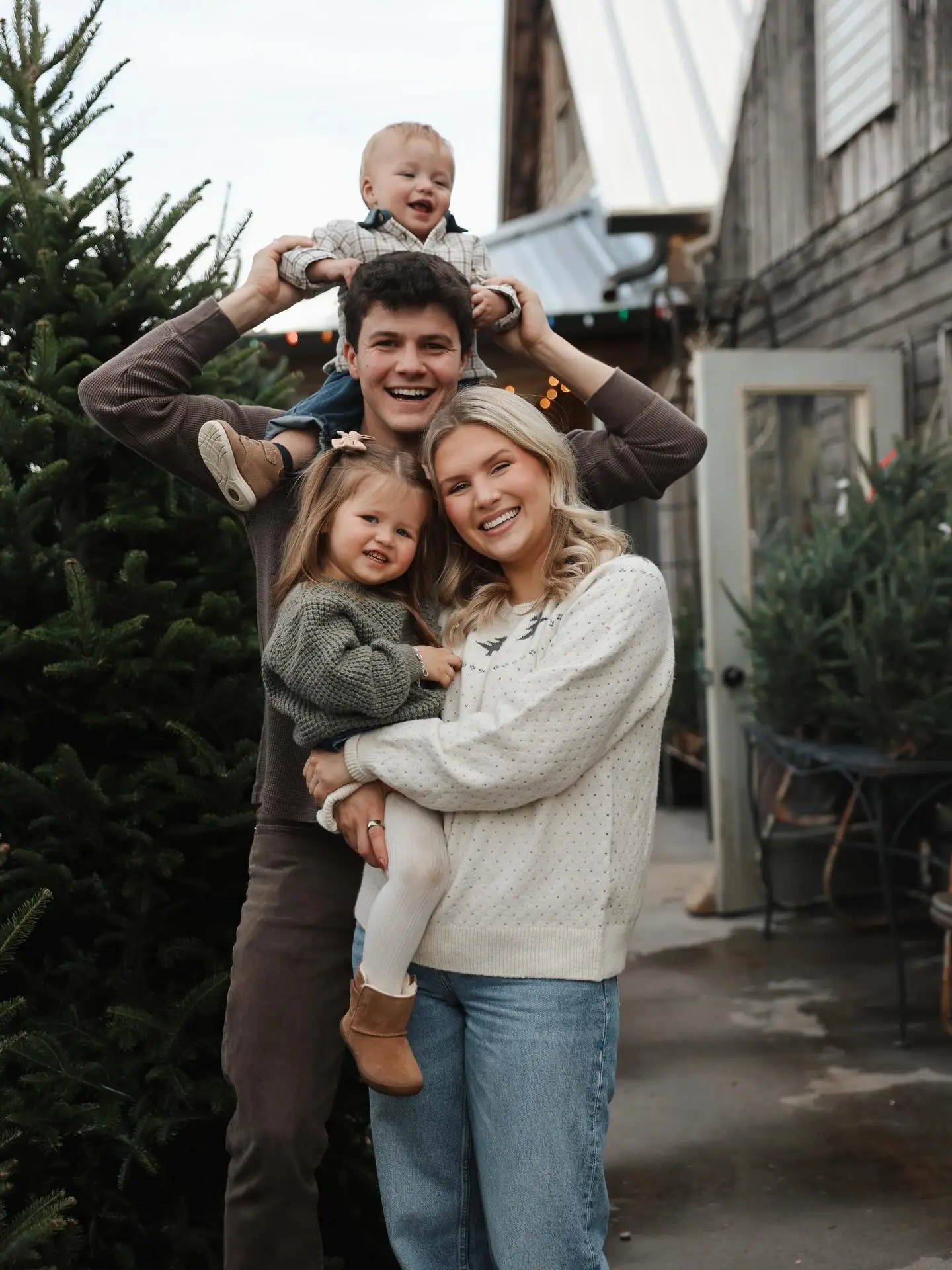 A family of four, including a man with a baby on his shoulders, a woman holding a young girl, all smiling while standing outdoors next to a Christmas tree.