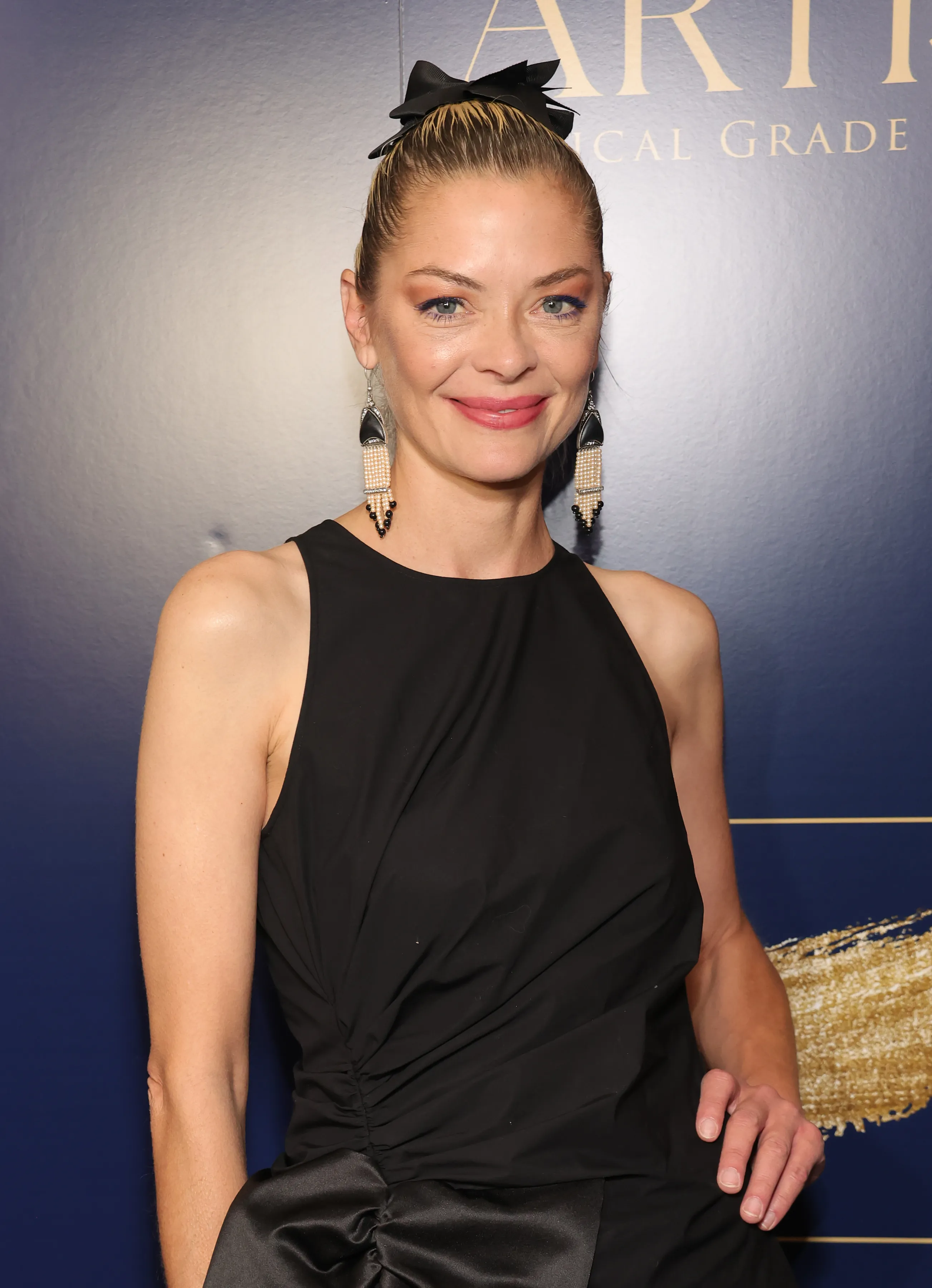 Jaime King poses in a black dress with a bow in her hair at the Artisa Skincare Launch Party.