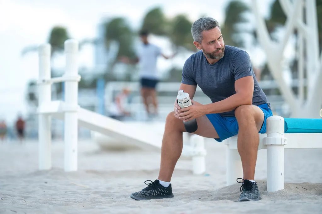 A man in athletic wear rests on a bench on the beach, holding a water bottle.