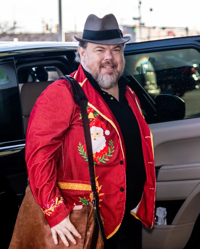 Devin Ratray wearing a red festive jacket with Santa Claus design, black shirt, and a brown fedora.