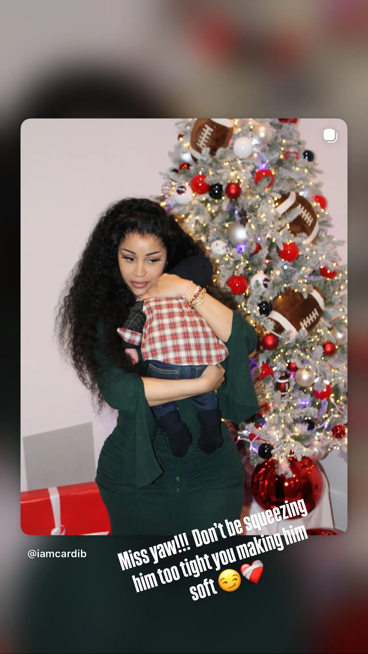 Cardi B holding a baby in front of a Christmas tree decorated with footballs.