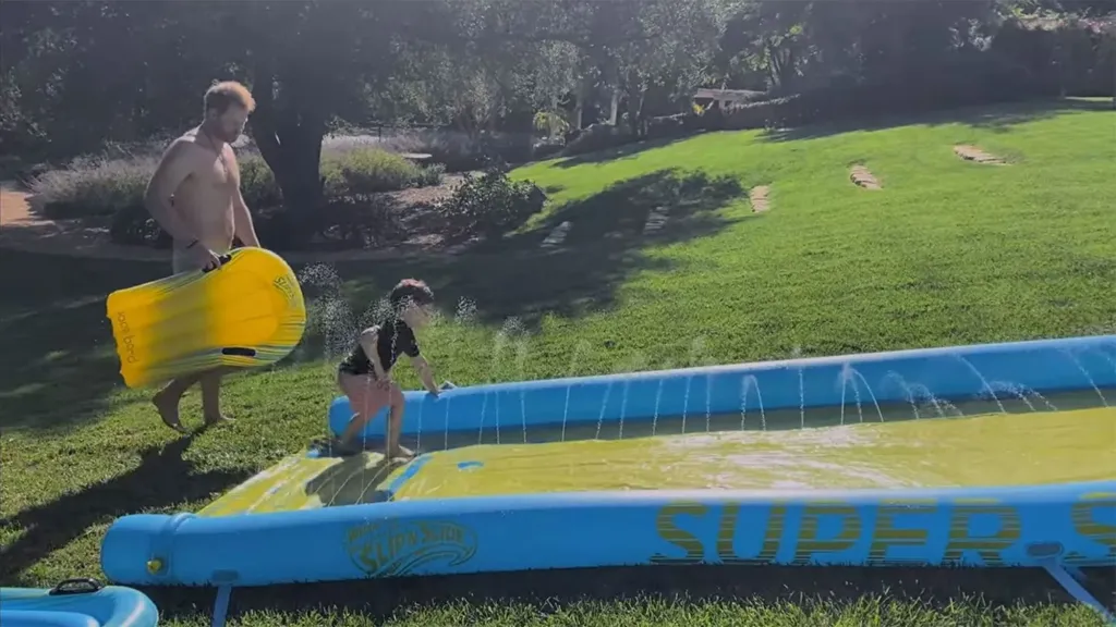 Prince Harry, in swim shorts, stands with Prince Archie, in a black t-shirt and pink shorts.