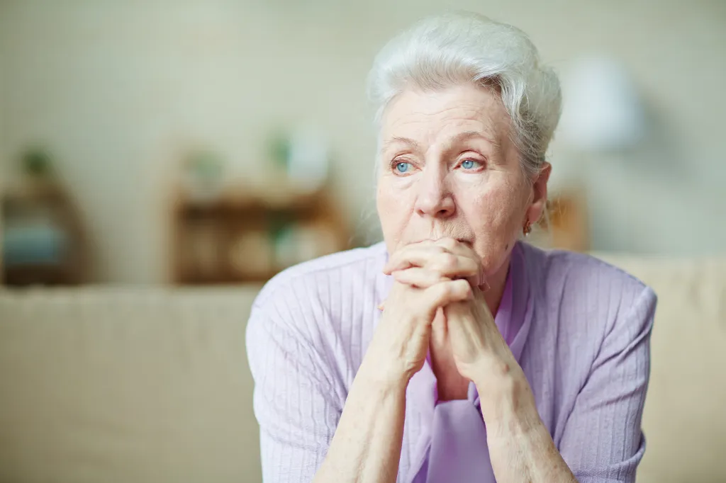 Elderly woman looking pensive or sad, with her hands clasped under her chin.