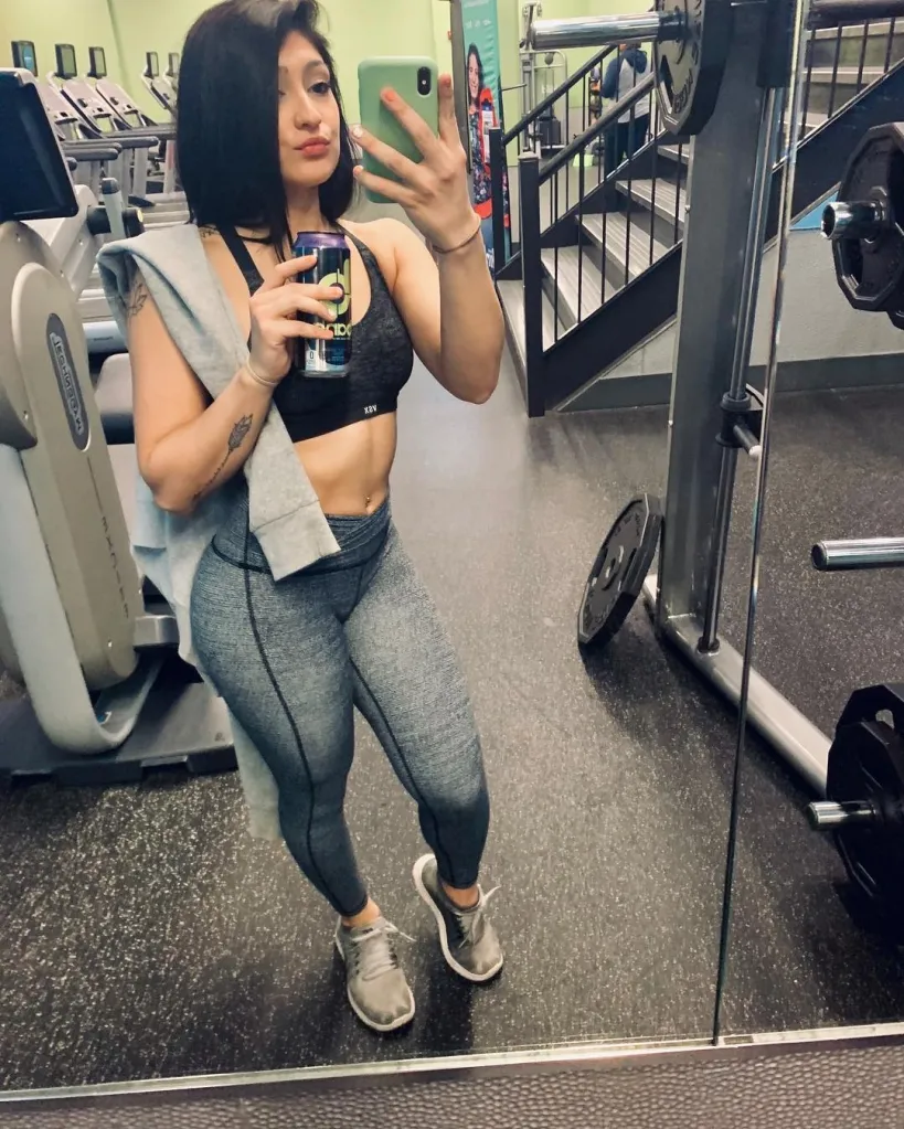 Woman in a sports bra and leggings takes a selfie in a gym mirror.