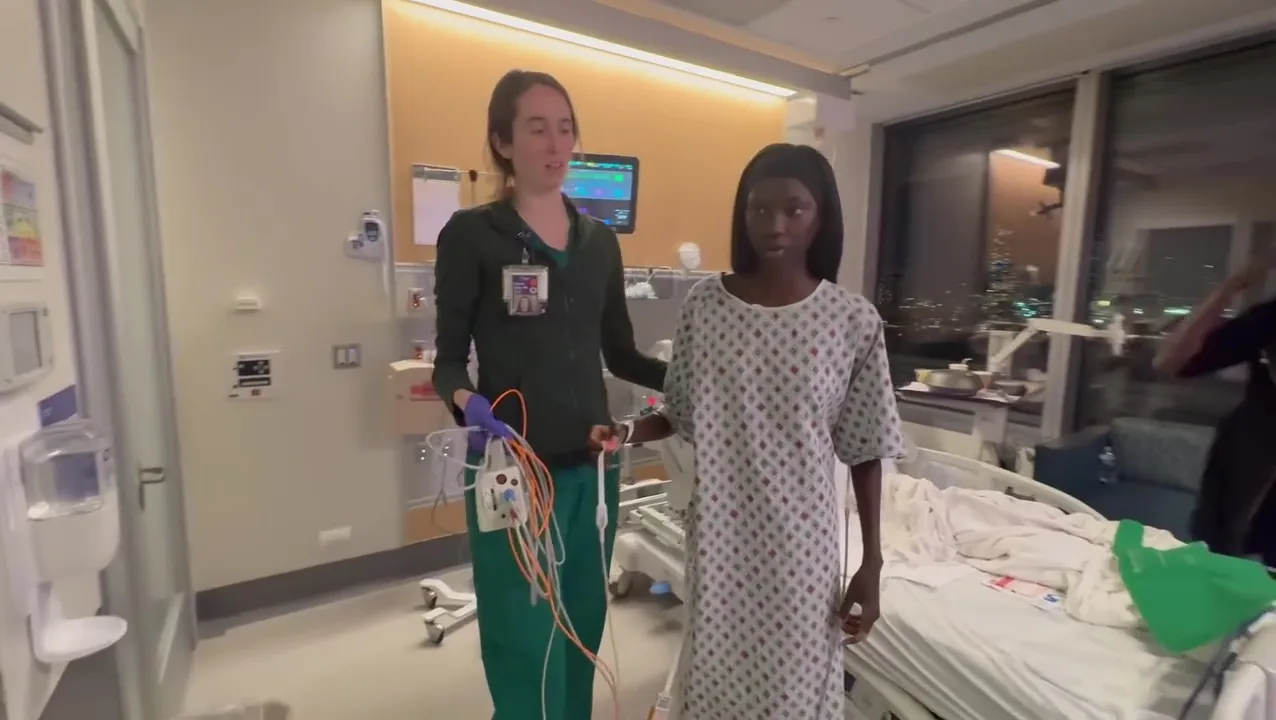 Anok Yai assisted by a nurse while walking in a hospital gown.