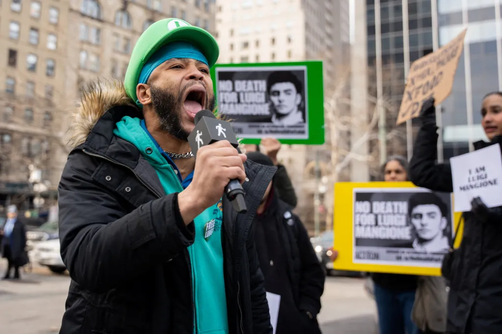 A man in a green Luigi hat and a blue hoodie yells into a microphone at a rally, with people behind him holding signs that read 