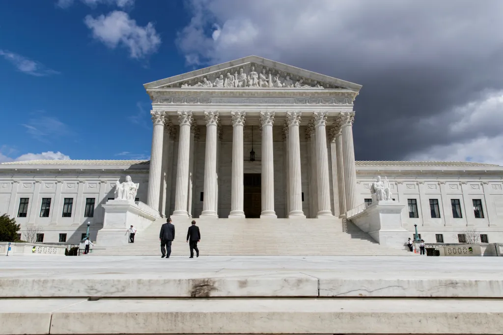 The U.S. Supreme Court building with a bright blue sky on the left and dark clouds on the right, as two men walk towards its entrance.