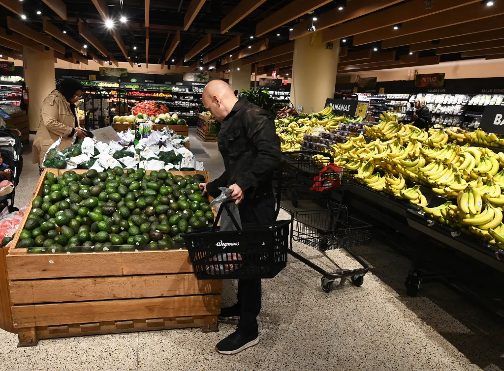 A bald man picking up an avocado from a bin, with bananas displayed to his right, in a Wegmans grocery store.