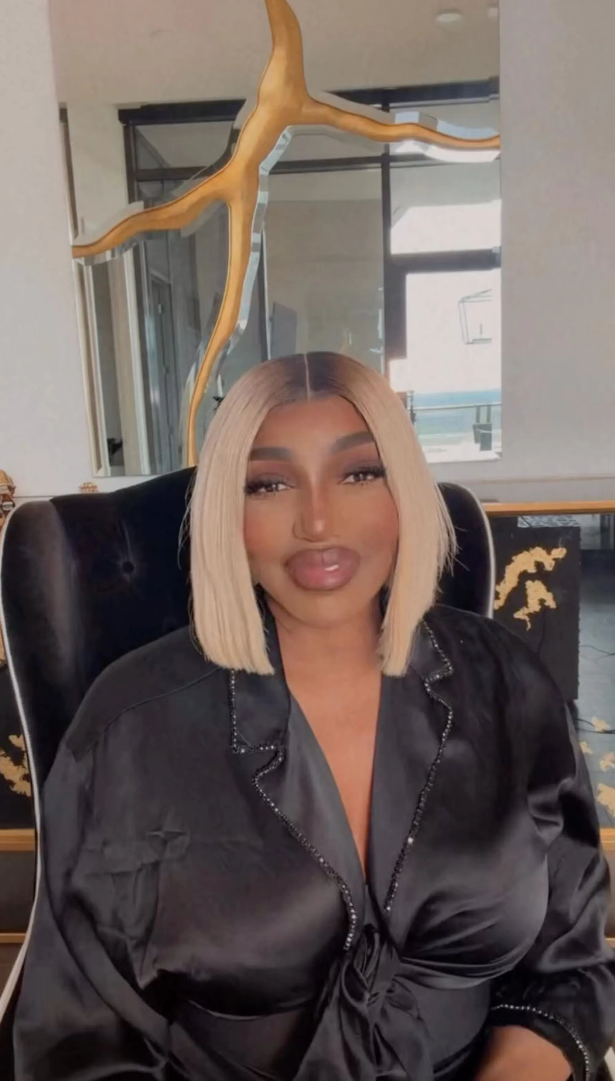 A woman with blonde hair, full lips, and a black satin top sitting in a black chair.