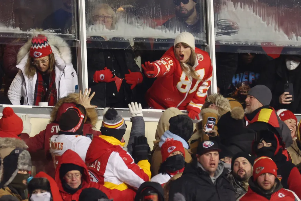 Taylor Swift in a red Chiefs jacket, white beanie, and red gloves, leans out of a stadium suite to celebrate with fans below.