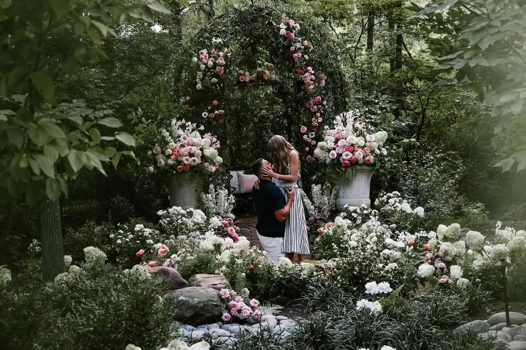 Man on one knee proposing to a woman in a lush garden with abundant pink and white flowers.