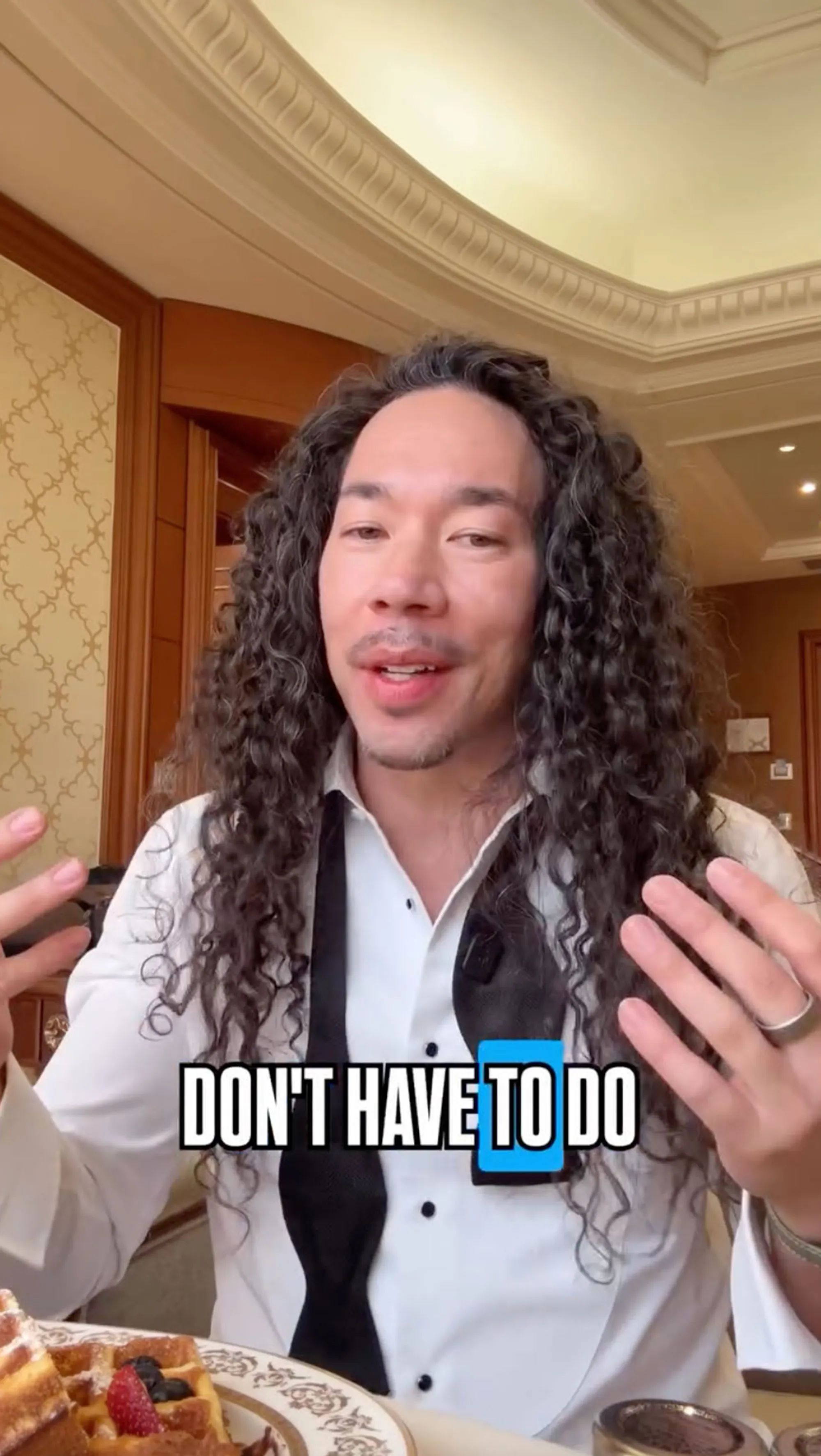Cole Walliser, with long curly hair, wearing a white shirt and black bowtie, speaks to the camera about Jennifer Lopez's shoot.