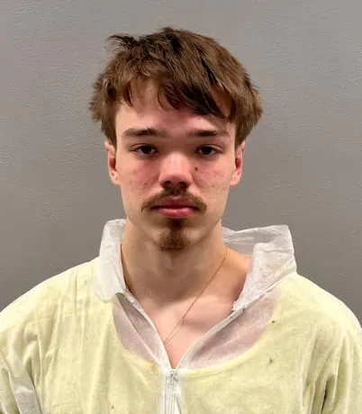 Mugshot of a young man with a mustache and goatee, wearing a white zip-up jumpsuit.